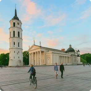 Dviratininkas ir du pėstieji juda per erdvią, atvirą aikštę su aukšta varpine ir dideliu baltu neoklasicistiniu pastatu su kolonomis fone. Saulei leidžiantis dangus yra mėlynų ir pastelinių spalvų mišinys.