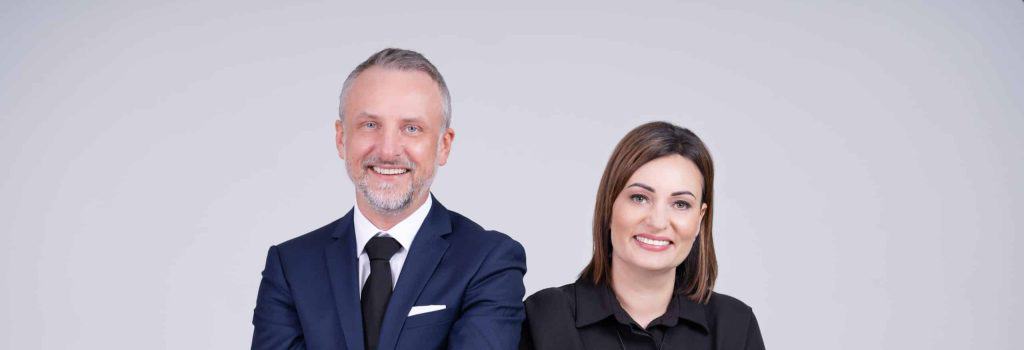 Vyras ir moteris šypsosi į kamerą. Vyras vilki tamsiai mėlyną kostiumą ir dėvi kaklaraištį, o moteris – juodą viršutinę dalį. Jie stovi paprastame šviesiai pilkame fone.