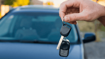 Ranka laiko porą automobilio raktelių su nuotolinio valdymo pulteliu priešais stovinčią transporto priemonę. Fone rodoma nefokusuota gatvės scena.