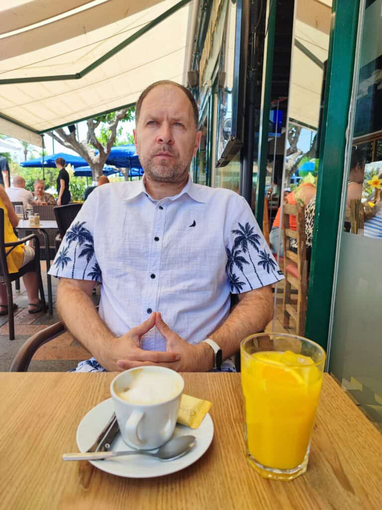 Vyras baltais marškiniais su palmių raštais sėdi prie lauko kavinės staliuko, priešais save turi puodelį kavos ir stiklinę apelsinų sulčių. Kiti žmonės sėdi aplink jį po skėčiais.