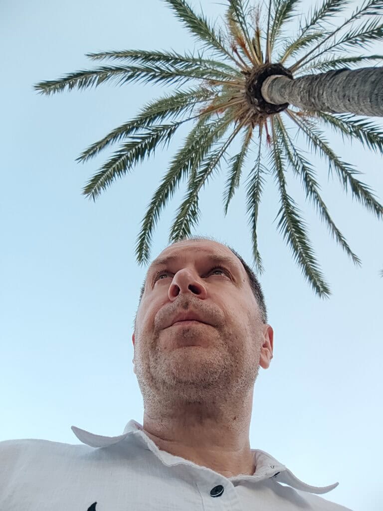 Vyras baltais marškiniais žvelgia į tolį, nufotografuotas nedideliu kampu, virš jo - aukšta palmė ir giedras mėlynas dangus.
