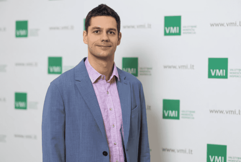 Vyras su šviesiai mėlynu švarkeliu ir rožiniais marškiniais stovi priešais baltą foną su žaliais VMI logotipais ir svetainės tekstu. Jis šypsosi ir žiūri į kamerą. | Sąskaita123 Vyras su šviesiai mėlynu švarkeliu ir rožiniais marškiniais stovi priešais baltą foną su žaliais VMI logotipais ir svetainės tekstu. Jis šypsosi ir žiūri į kamerą.