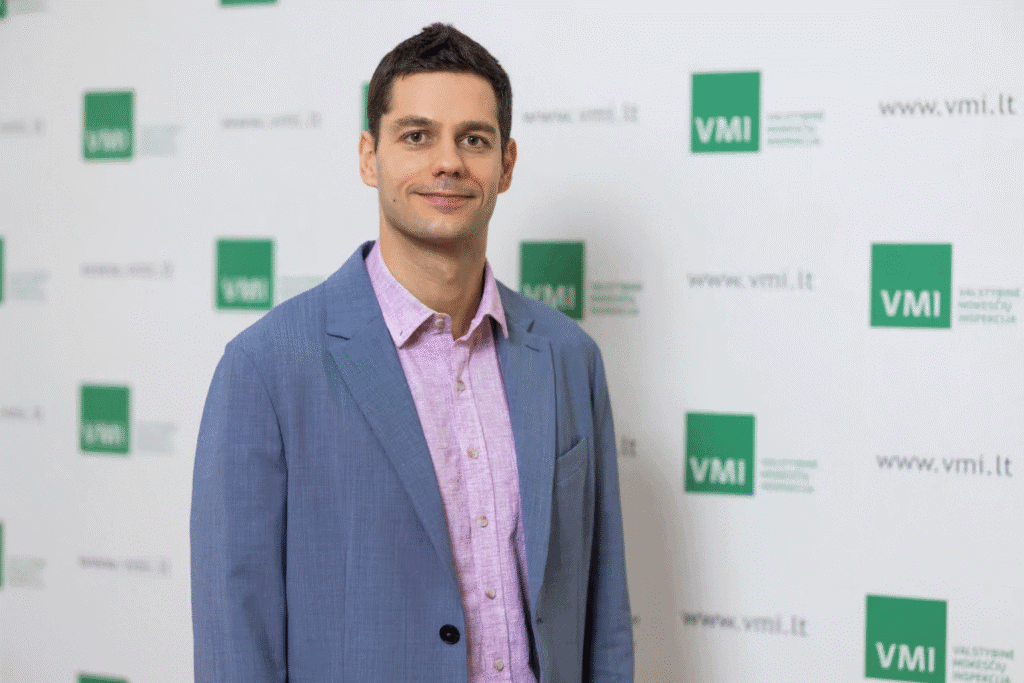 Vyras su mėlynu švarkeliu ir šviesiai violetiniais marškiniais stovi priešais foną su keliais žaliais VMI logotipais ir tekstais, pozuoja ir šypsosi į kamerą.