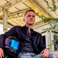 Jaunas vyras juoda striuke sėdi lauke prie pastato su metaliniu stogu ir laiko motociklininko šalmą su spalvotu skydeliu. Fone matomi žali lapai ir dalis statinio.