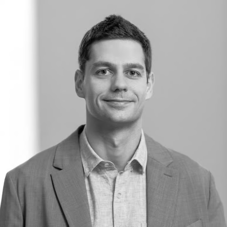 Trumpais tamsiais plaukais vilkintis vyras, vilkintis šviesius marškinius ir švarką, šiek tiek šypsosi žiūrėdamas į kamerą. Nuotrauka nespalvota, vienspalvė, vienspalviame fone.